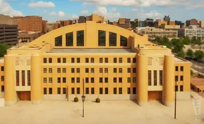 An aerial view of a large, tan building with a distinctive arched roof and numerous windows, set against a backdrop of a city skyline under a cloudy sky.