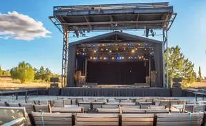 Outdoor amphitheater stage with rows of empty wooden seats facing the stage under a bright blue sky.