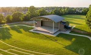 Outdoor amphitheater stage with covered seating and tiered grassy lawn in a park setting during sunset.