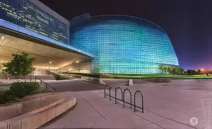 Modern glass building with blue-green lights at night, in front of a plaza with bike racks.