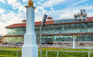 A white column topped with a gold ball stands in front of a modern grandstand at a racetrack under a cloudy sky.