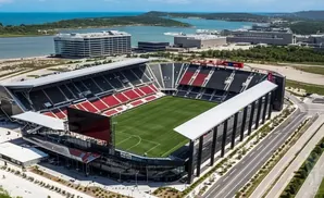 Aerial view of a modern soccer stadium with red and black seats, overlooking a body of water and distant buildings.