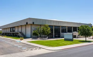 A large, modern concrete building with glass windows and a white roof under a clear blue sky.