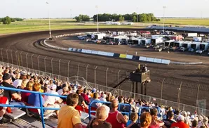 Fans watch a dirt track race at a stadium, with cars lined up in the pit area.