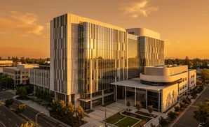 Modern glass and concrete buildings reflect golden sunset light, surrounded by trees and a street.