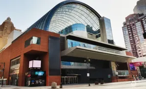 A modern building with a glass dome roof and an orange facade, surrounded by tall skyscrapers under a clear sky.