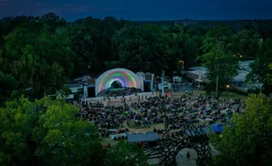 An aerial view shows an outdoor concert with a rainbow-lit stage and a large audience seated on the grass.