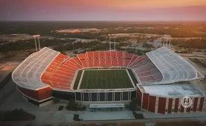 An aerial view of an empty football stadium with red seats and a green field during sunset.