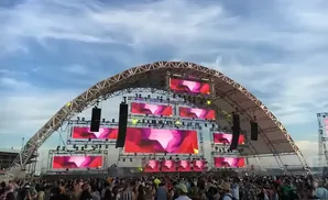 Large outdoor stage with multiple screens displaying abstract pink and purple visuals, surrounded by a crowd of people.