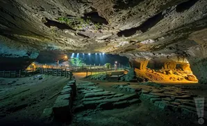 A vast cave with stone pathways and wooden fences, illuminated by lights at the opening.
