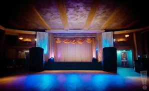 An empty stage in a theater with a curtain and stage lights, casting blue and purple hues on the floor.