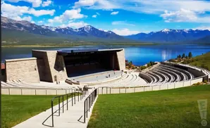 Outdoor amphitheater with stage overlooking a lake and snow-capped mountains under a blue, cloudy sky.