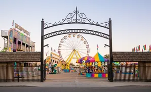 An amusement park entrance with a Ferris wheel, rides, and game booths under a clear sky.