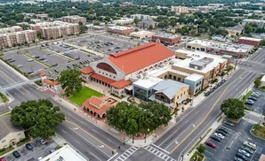 Aerial view of a downtown area with large buildings, a red-roofed structure, a parking lot, and trees.