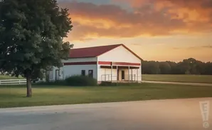 A white building with a red roof and trim sits beside a large tree under a colorful sunset sky.