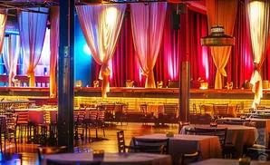 A dimly lit event space with tables set for dining, draped curtains, and stage lighting.