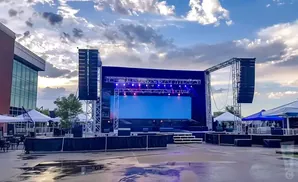 Outdoor stage with large screen, speakers, tents, and a cloudy sky.