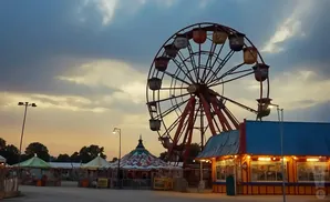 An amusement park with a Ferris wheel, circus tents, and food stands under a cloudy twilight sky.