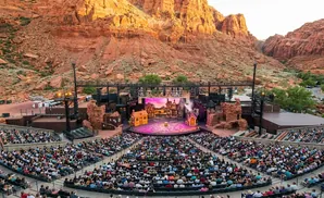 Outdoor theater in a canyon with a stage set with houses and a large audience seated in tiered rows.