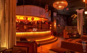 Interior of a dimly lit bar with orange lighting, a curved bar, plush seating, and ornate chandeliers.