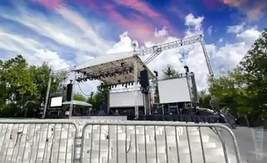 An outdoor concert stage with empty white seats in the foreground under a colorful sky.