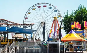 A Ferris wheel and roller coaster at a sunny amusement park with colorful tents and flags.