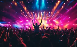 A concert crowd with hands in the air and a performer on stage under colorful lights.