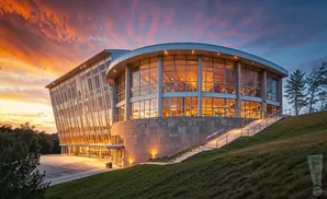 Modern glass building with a curved facade and lit interior, at sunset with colorful sky.