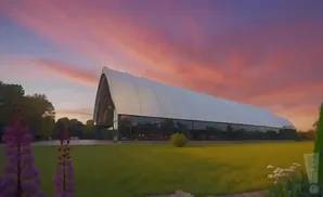 A large white arched building with glass windows sits on a grassy lawn under a vibrant pink and purple sunset sky.