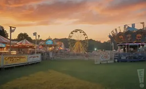 Amusement park rides, including a Ferris wheel and carousel, are set up on a grassy field under a colorful sunset sky.