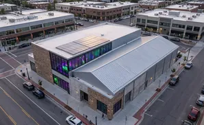 An aerial view of a modern brick and glass building with solar panels on the roof, parked cars, and city streets.