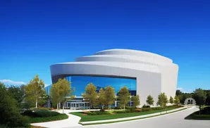 Modern building with wavy roof and large blue glass windows, surrounded by trees and green lawns.