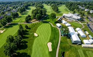 Aerial view of a golf course with fairways, greens, sand traps, trees, houses, tents, and a clubhouse.