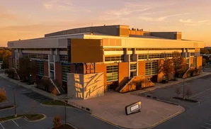 An arena with a large screen stands at sunset, with a parking lot and trees.