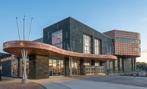 Modern building with dark stone exterior, copper canopy, and large windows under a blue sky.