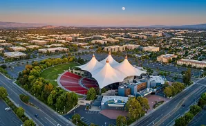 Aerial view of a modern amphitheater with a large white tent roof and surrounding cityscape at sunset.