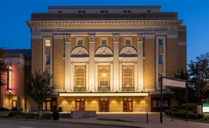 A grand brick theater at dusk, lit from within and by external lighting, with trees and signage outside.