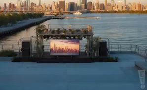 Outdoor stage with a large screen showing a city skyline at sunset, overlooking a body of water.