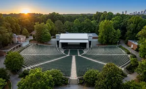 Outdoor amphitheater with tiered seating, stage, and surrounding trees, with a city skyline in the distance at sunset.