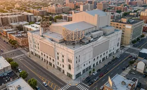 Aerial view of a large white building with a distinctive metal structure on its roof, surrounded by city streets and buildings.