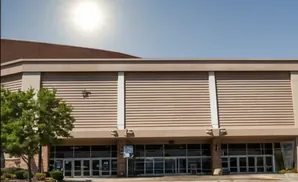 A large building with a tan, horizontal-slatted facade and a brown roof under a bright sun.