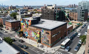 An aerial view of a city street with a large mural on a brick building, cars, and construction cranes.