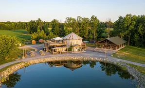 Aerial view of a rustic venue with a silo building, covered pavilion, and pond, surrounded by trees and fields.