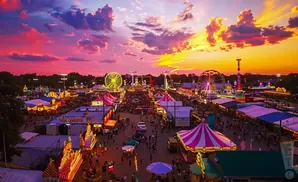 A vibrant fairground at sunset with Ferris wheels, tents, and crowds under a dramatic pink and orange sky.