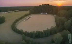 An aerial drone view of a dirt racetrack surrounded by trees at sunset.