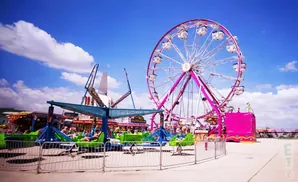 A pink Ferris wheel and a thrill ride stand under a bright blue sky with scattered clouds at a carnival.