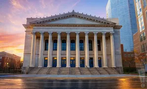 A classical building with large columns and blue doors is reflected on a wet street at sunset.