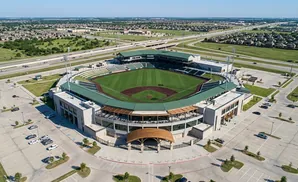 Aerial view of a baseball stadium with surrounding parking lots and residential areas under a clear blue sky.