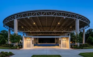 An outdoor amphitheater with a circular, metal roof supported by large concrete pillars. Blue seats are visible on the ground.