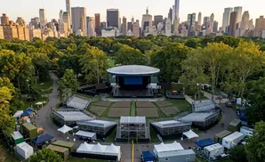 Aerial view of an outdoor amphitheater with tiered seating, a stage, and a city skyline in the background.
